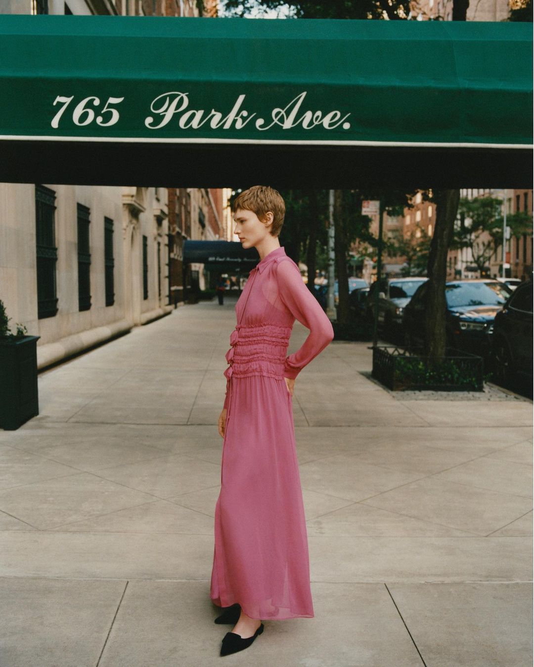Woman in a pink dress stands on a sidewalk in front of a building at 765 Park Ave. in an urban setting, captured in natural light.
