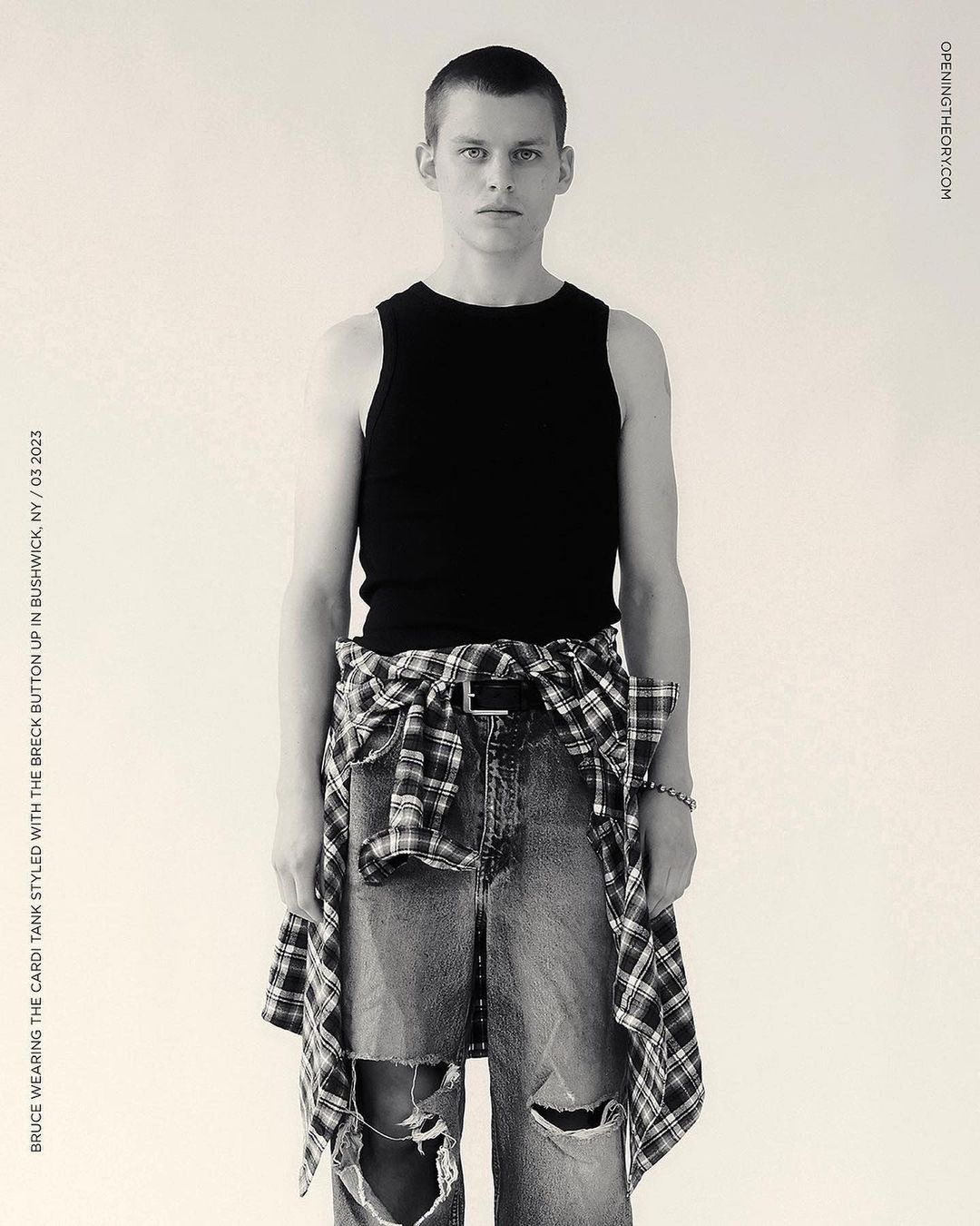 Full shot of a young adult man in a black tank top and distressed jeans, styled with a plaid shirt tied around his waist, against a plain backdrop.