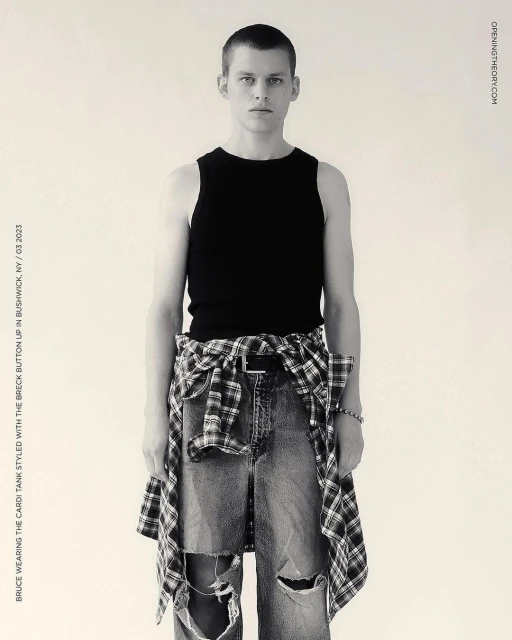 Full shot of a young adult man in a black tank top and distressed jeans, styled with a plaid shirt tied around his waist, against a plain backdrop.