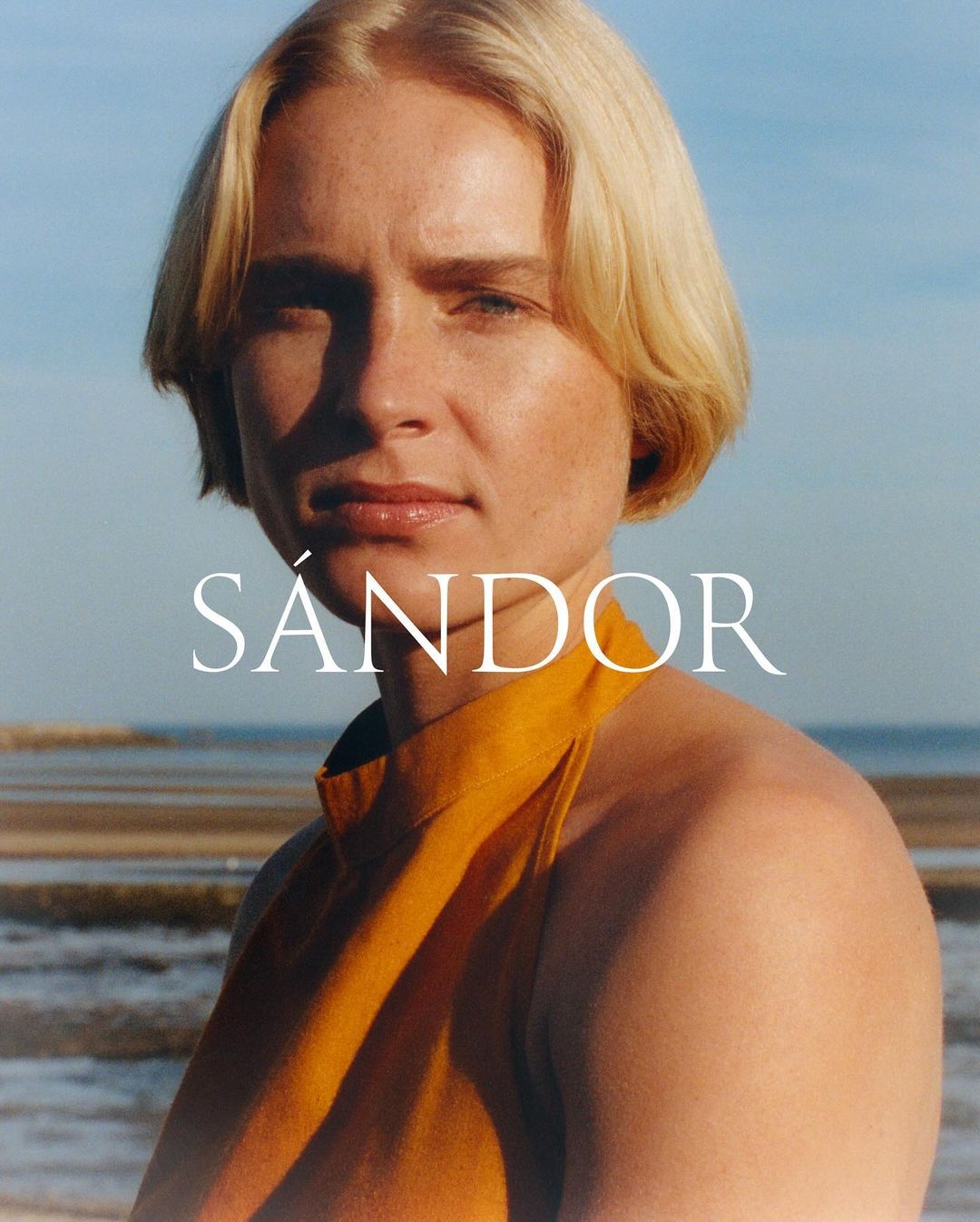 Portrait of a blonde woman wearing a yellow halter top outdoors against a sandy beach and ocean background.