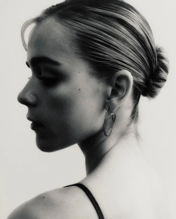 Monochrome portrait of a fair-skinned woman looking over her shoulder wearing sculptural earrings in a minimalist studio setting.