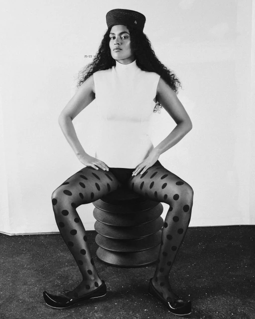 A woman with curly hair sits on a circular, stacked object in a sleeveless turtleneck and patterned tights in a studio setting.