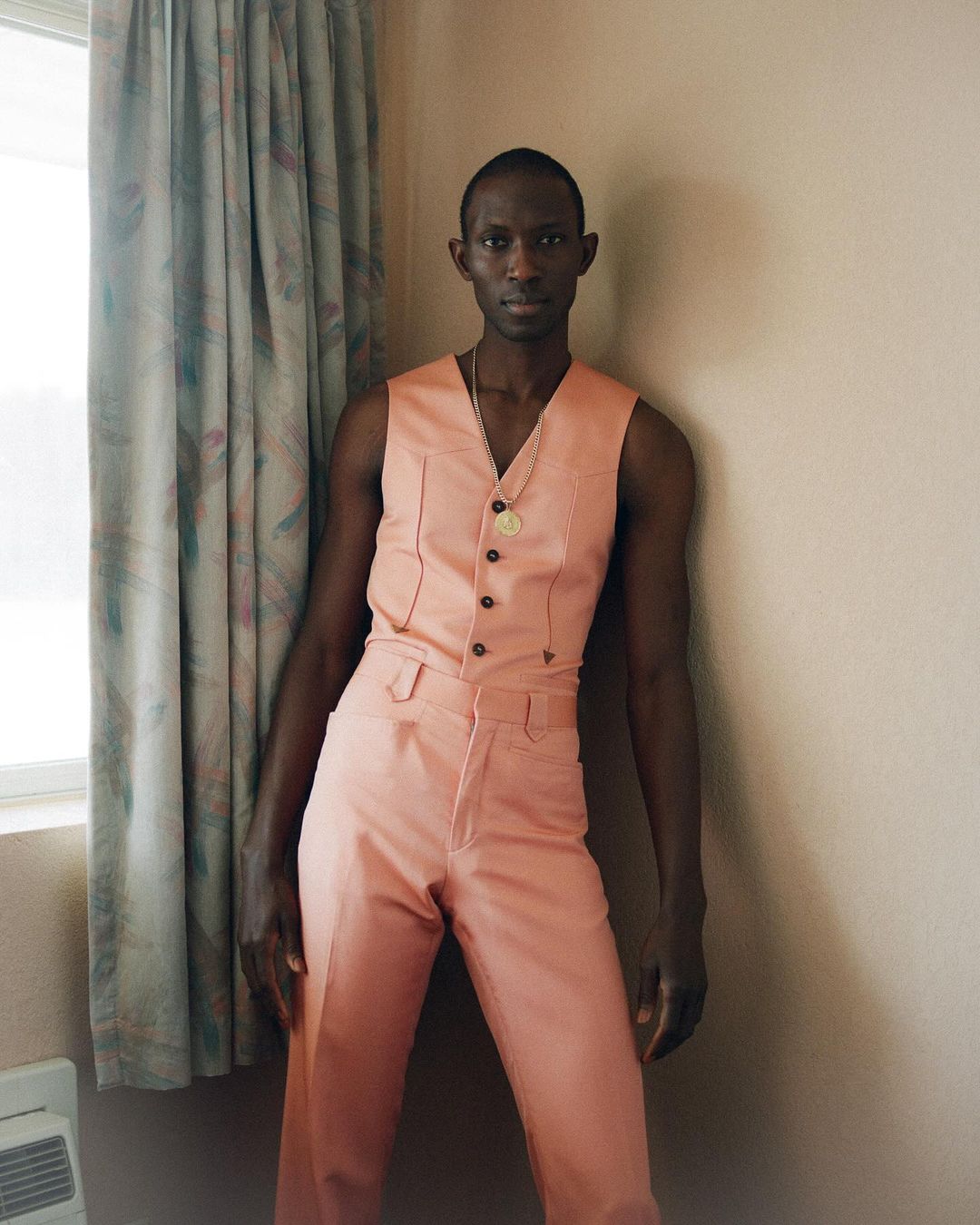 A slender Black man in a peach-colored suit vest and trousers stands indoors, leaning against a peach wall next to a patterned curtain.