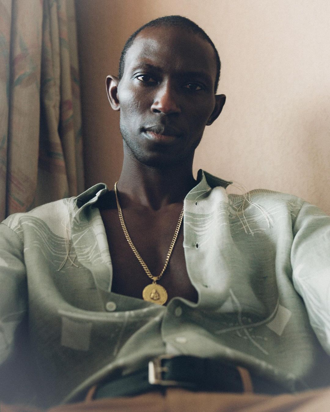 Portrait of a Black man in casual patterned shirt and gold chain indoors. He is seated and looking directly at the camera.