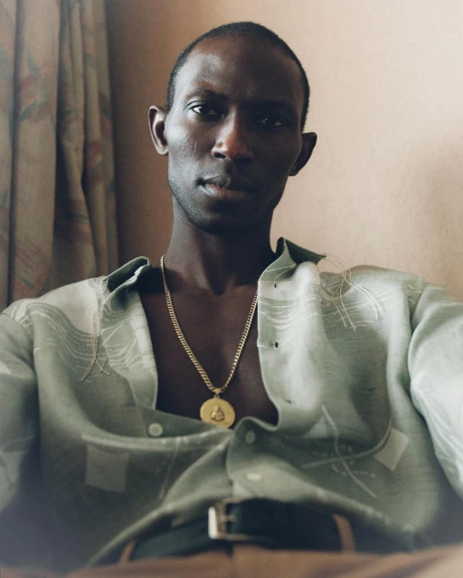 Portrait of a Black man in casual patterned shirt and gold chain indoors. He is seated and looking directly at the camera.