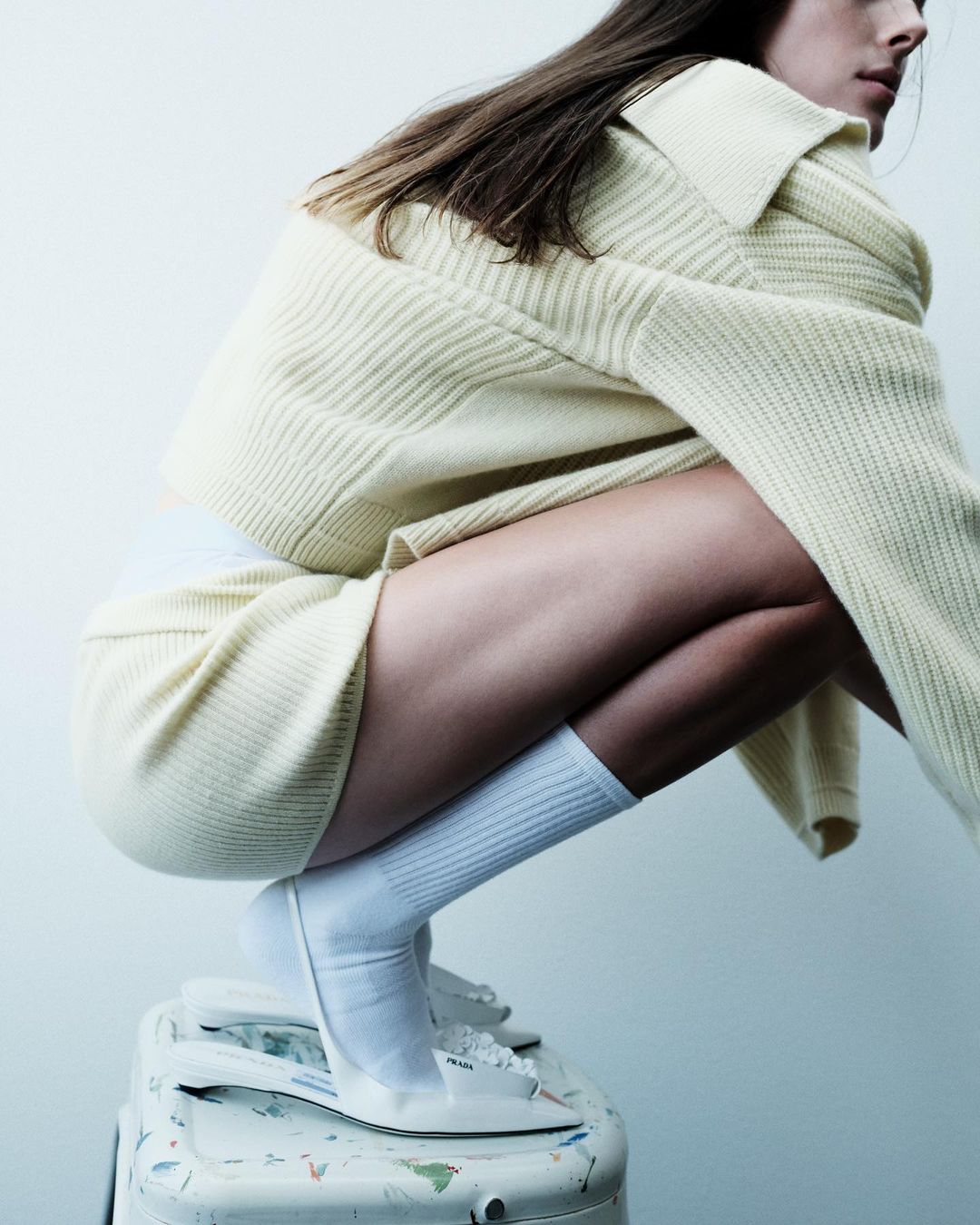 A fair-skinned young woman in a cream-colored knit sweater and shorts crouching on a paint-splattered stool in a minimalist setting.
