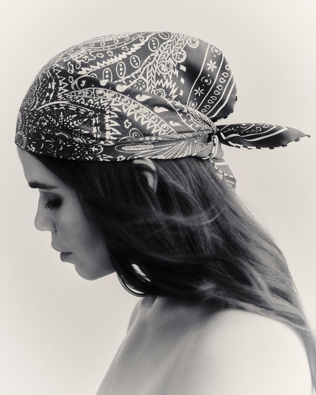 Black and white portrait of a woman wearing a paisley bandana tied on her head, with long hair, and looking down in a photo studio.