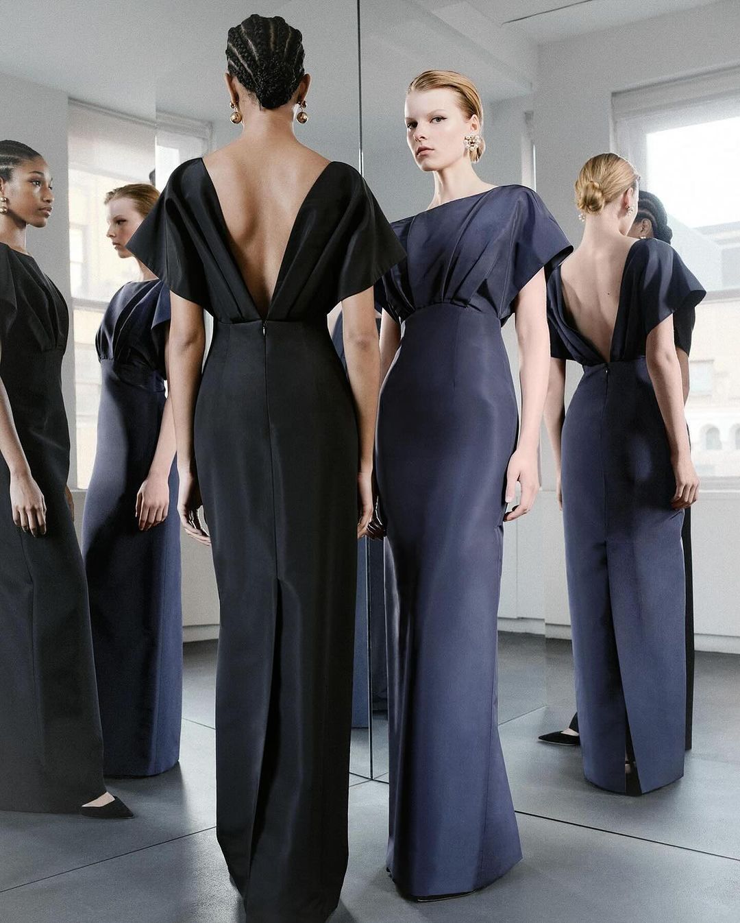 Elegant women in long gowns at a fashion show, captured indoors with soft lighting and a mirror creating multiple reflections.