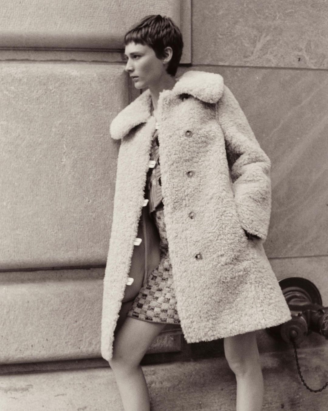 A woman in a white coat stands against a concrete wall with a fire hydrant, casting a vintage, fashion-focused, outdoor scene.