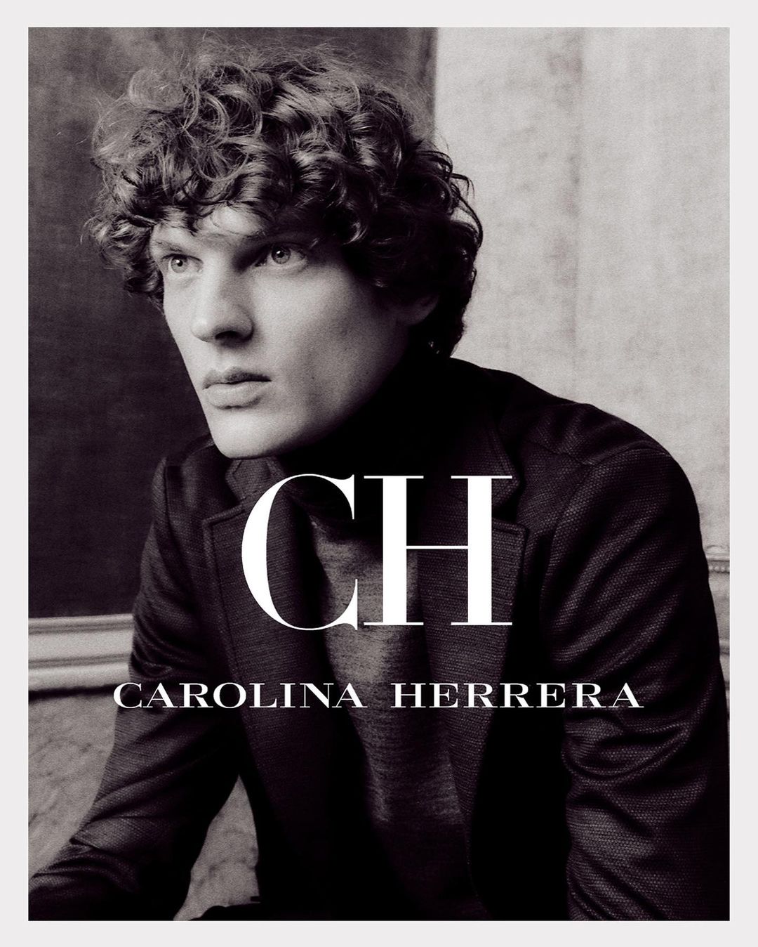 Black and white fashion-style portrait of a young adult male model with curly hair wearing dark jacket with Carolina Herrera logo.