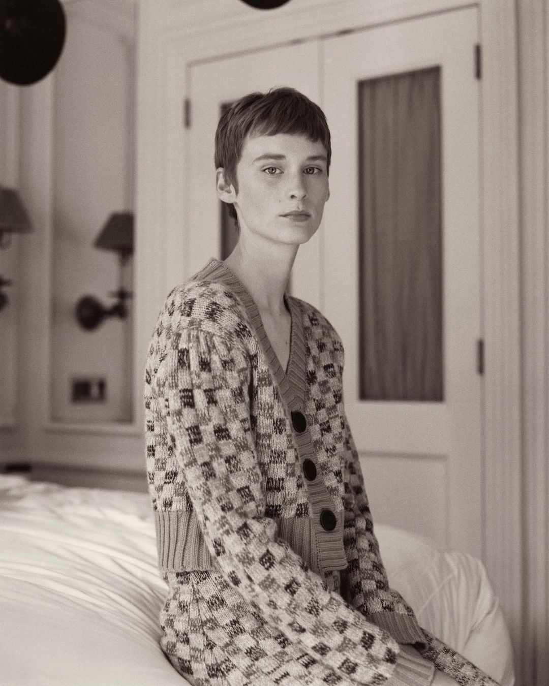 Adult female with short hair sits indoors on a bed. She wears a patterned knit cardigan with buttons, against a neutral interior setting.