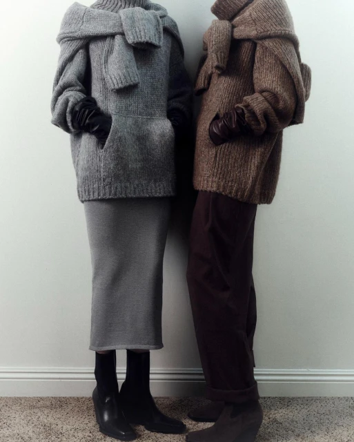 Two people wearing matching knitwear outfits in a studio setting; they have their hands in their pockets and are facing the camera.