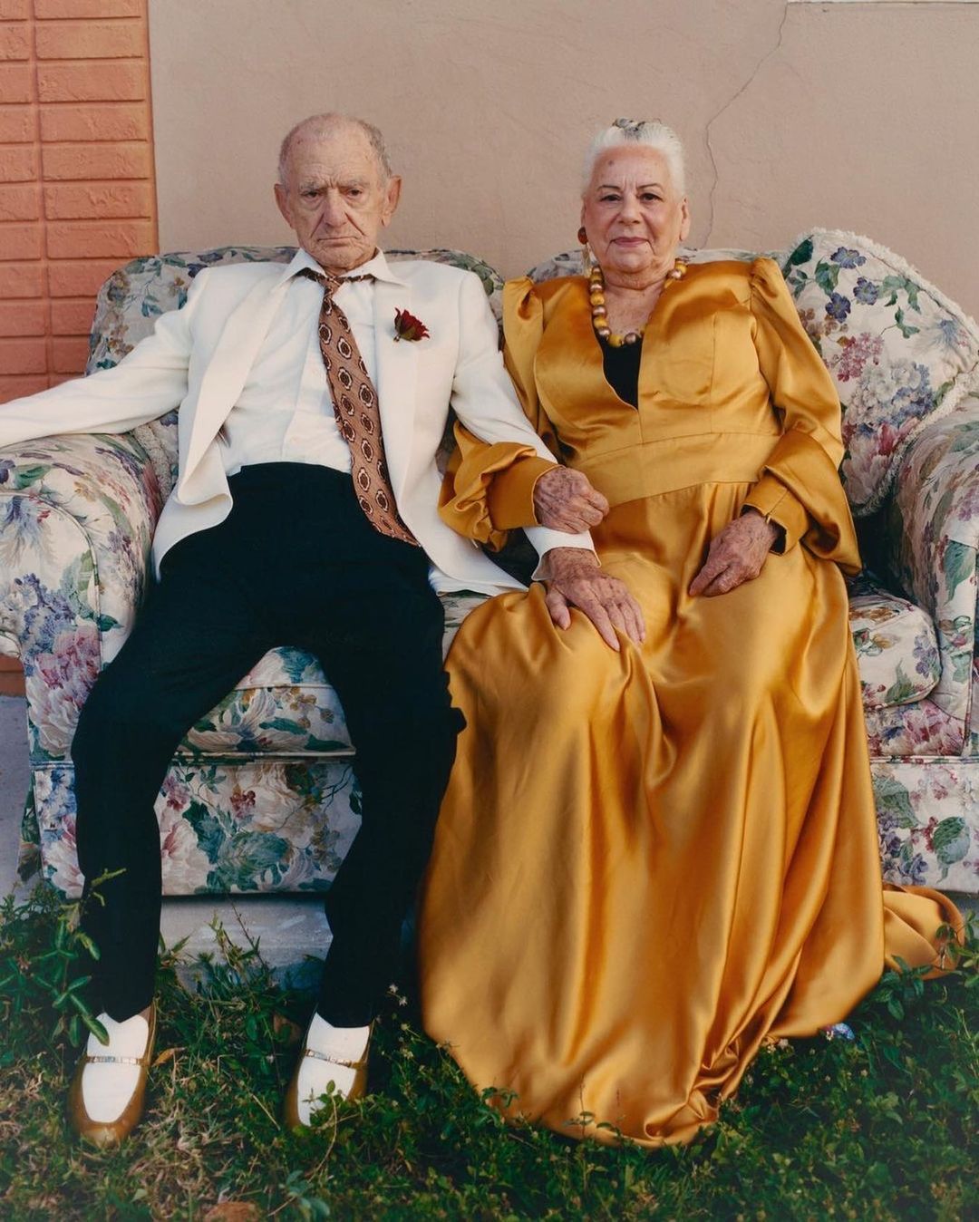 Elderly couple dressed formally poses on floral sofa outdoors. Older man in white suit, older woman in gold satin dress, holding hands.