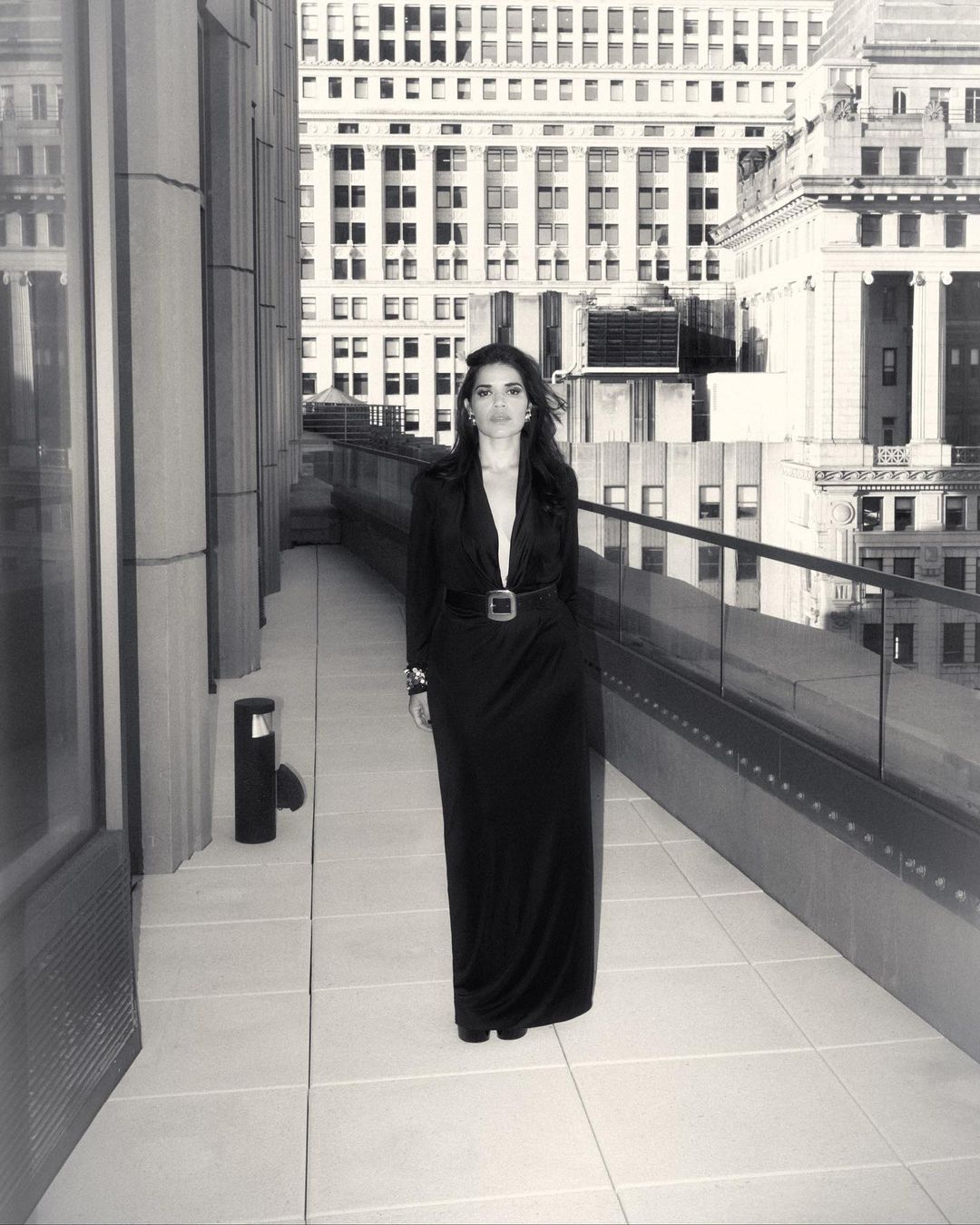 Woman in a black dress stands on a city terrace with tall buildings in the background in a black-and-white fashion shot.