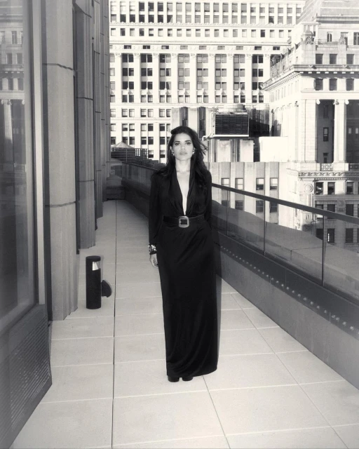 Woman in a black dress stands on a city terrace with tall buildings in the background in a black-and-white fashion shot.