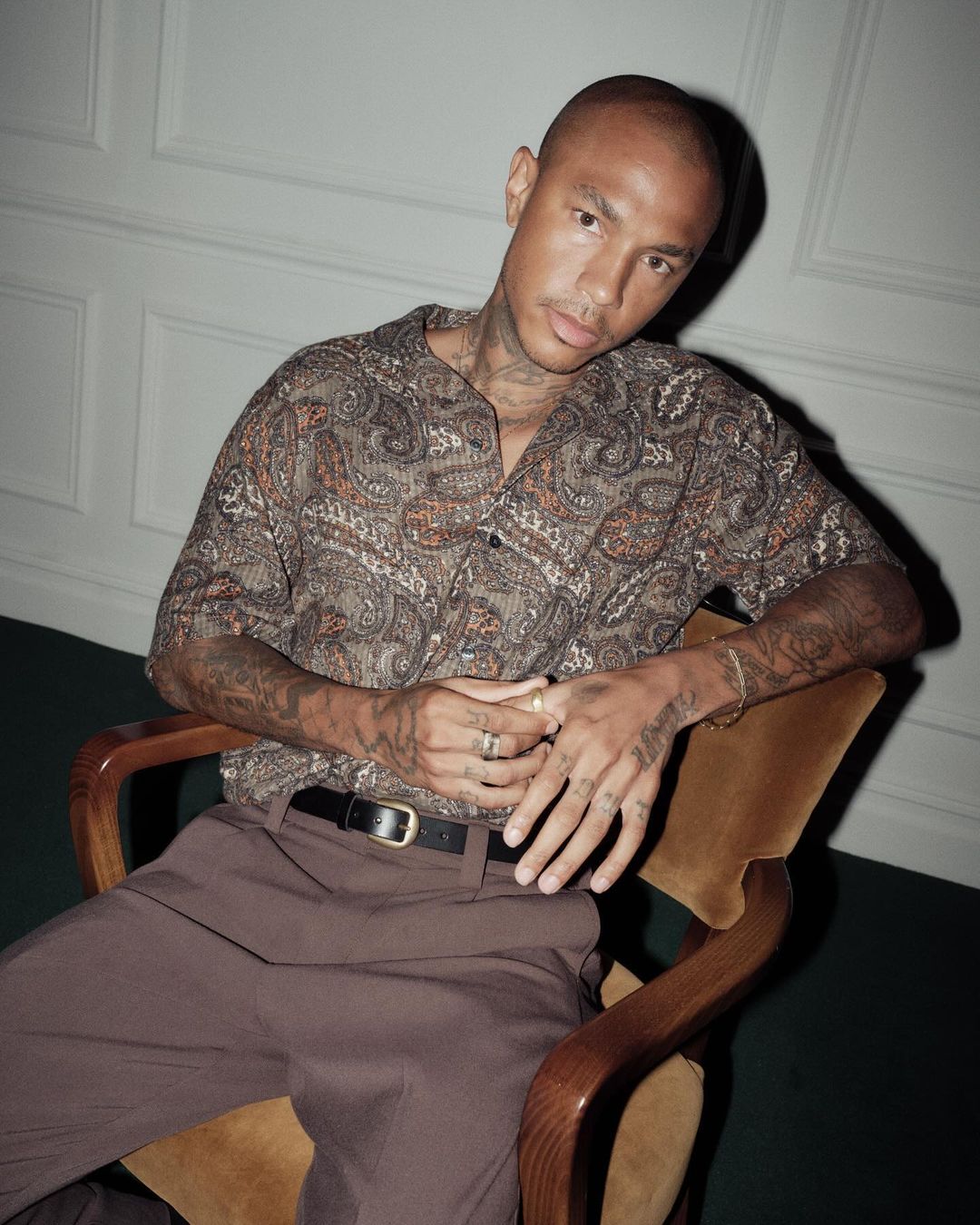 Stylish man with tattoos poses indoors in a patterned short-sleeved shirt, brown trousers, and a belt while sitting on a chair.