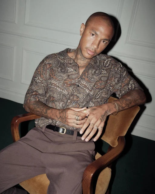 Stylish man with tattoos poses indoors in a patterned short-sleeved shirt, brown trousers, and a belt while sitting on a chair.