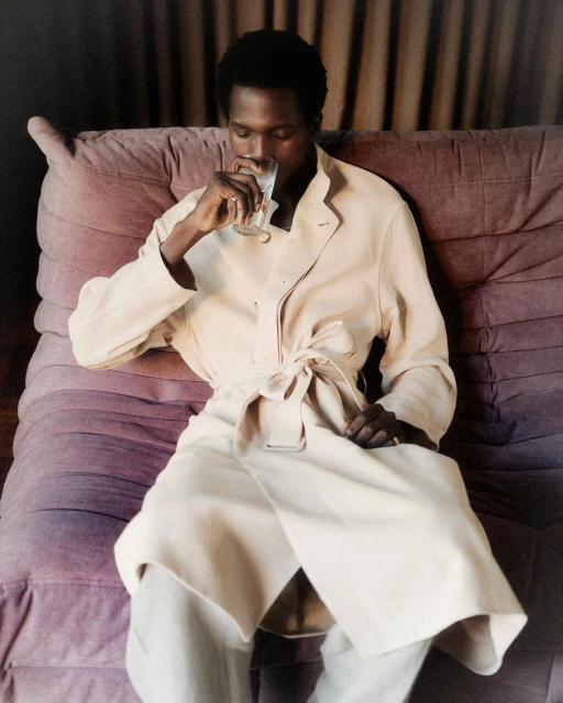 An adult Black man in a long beige coat lounges on a modern purple sofa, holding a glass of water in a comfortable, indoor setting.