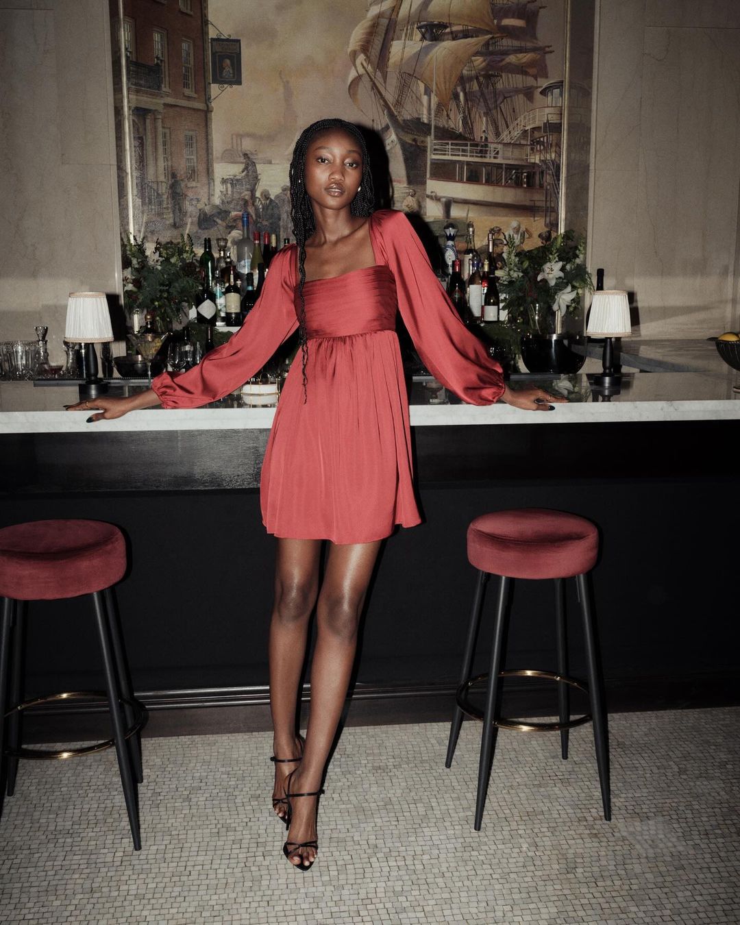 Elegant adult female in a terracotta-colored dress stands in front of a bar, showcasing fashion and style in an indoor environment.