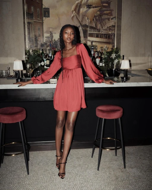 Elegant adult female in a terracotta-colored dress stands in front of a bar, showcasing fashion and style in an indoor environment.