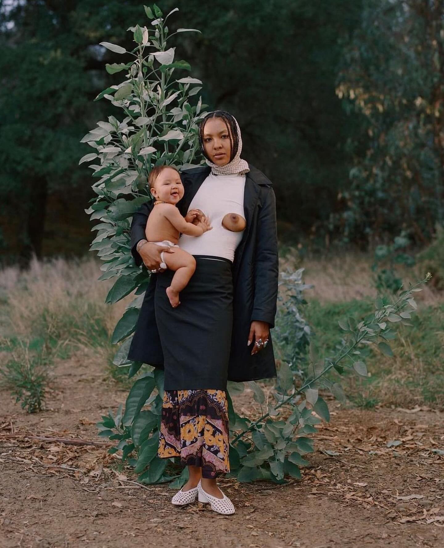 Woman wearing a black coat and patterned skirt holding a smiling baby outdoors before green trees.