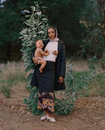 Woman wearing a black coat and patterned skirt holding a smiling baby outdoors before green trees.