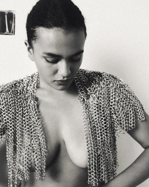Monochrome portrait of a woman wearing metal chainmail, shot in a photo studio with a plain backdrop and diffused, even lighting.