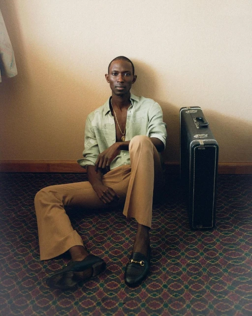A Black man sits on a patterned floor wearing casual attire with a suitcase beside him, creating a relaxed, indoor editorial portrait.