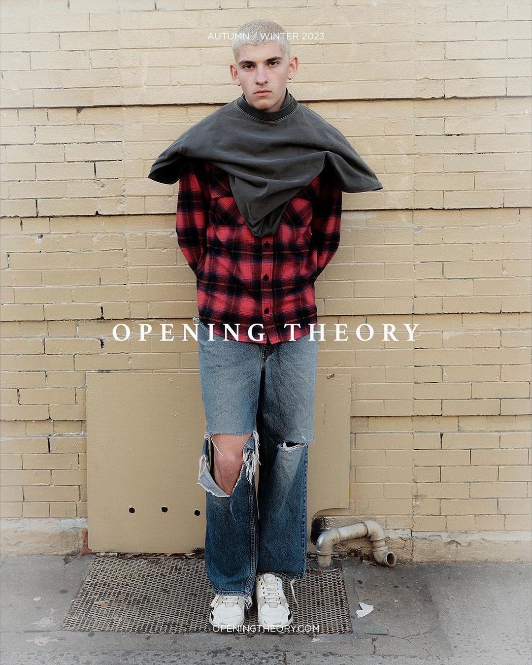 Fashion shot of a male model in tattered jeans, plaid shirt, and grey top, posing against a brick wall for Opening Theory Autumn/Winter 2023.