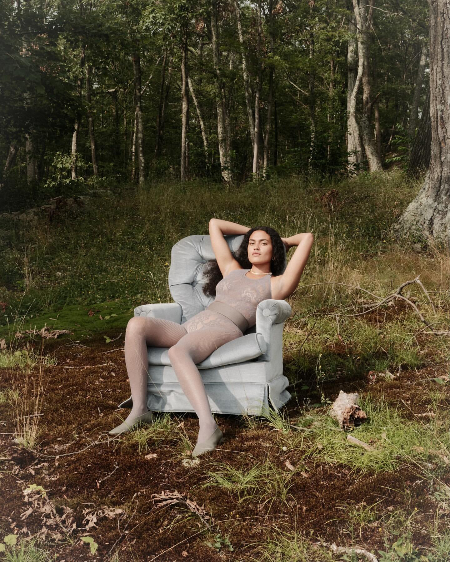 Woman in sheer lingerie and tights lounges in an armchair outdoors amid tall grass and trees in a lush forest landscape, natural lighting.