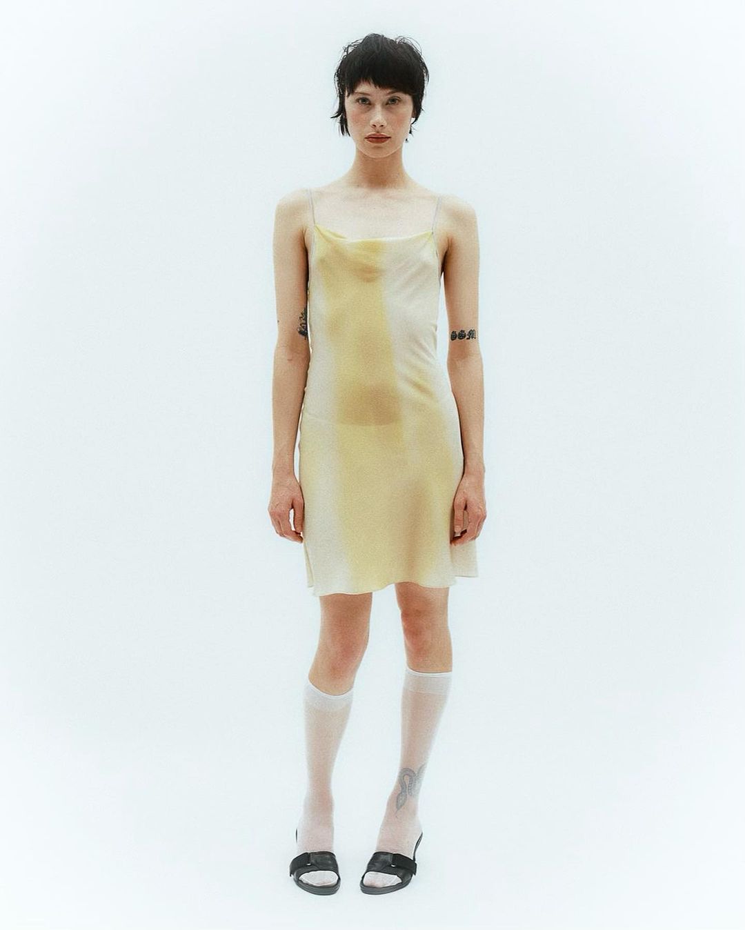 A slender woman in a pale yellow slip dress stands against a bright white backdrop, complemented by knee-high socks and black shoes.