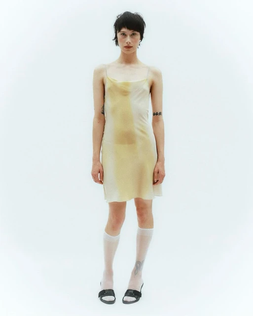 A slender woman in a pale yellow slip dress stands against a bright white backdrop, complemented by knee-high socks and black shoes.