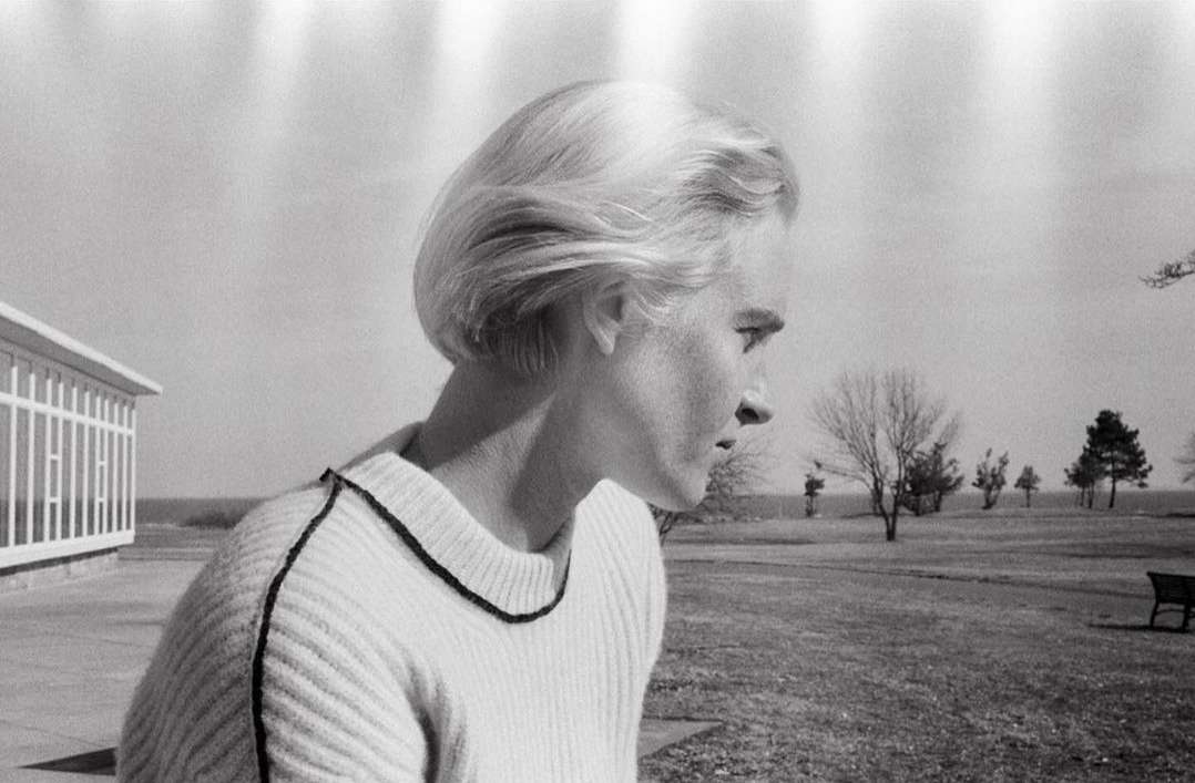 Monochrome portrait of a blonde woman in a sweater, looking right, with a building and landscape visible in the background.