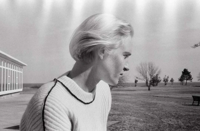 Monochrome portrait of a blonde woman in a sweater, looking right, with a building and landscape visible in the background.