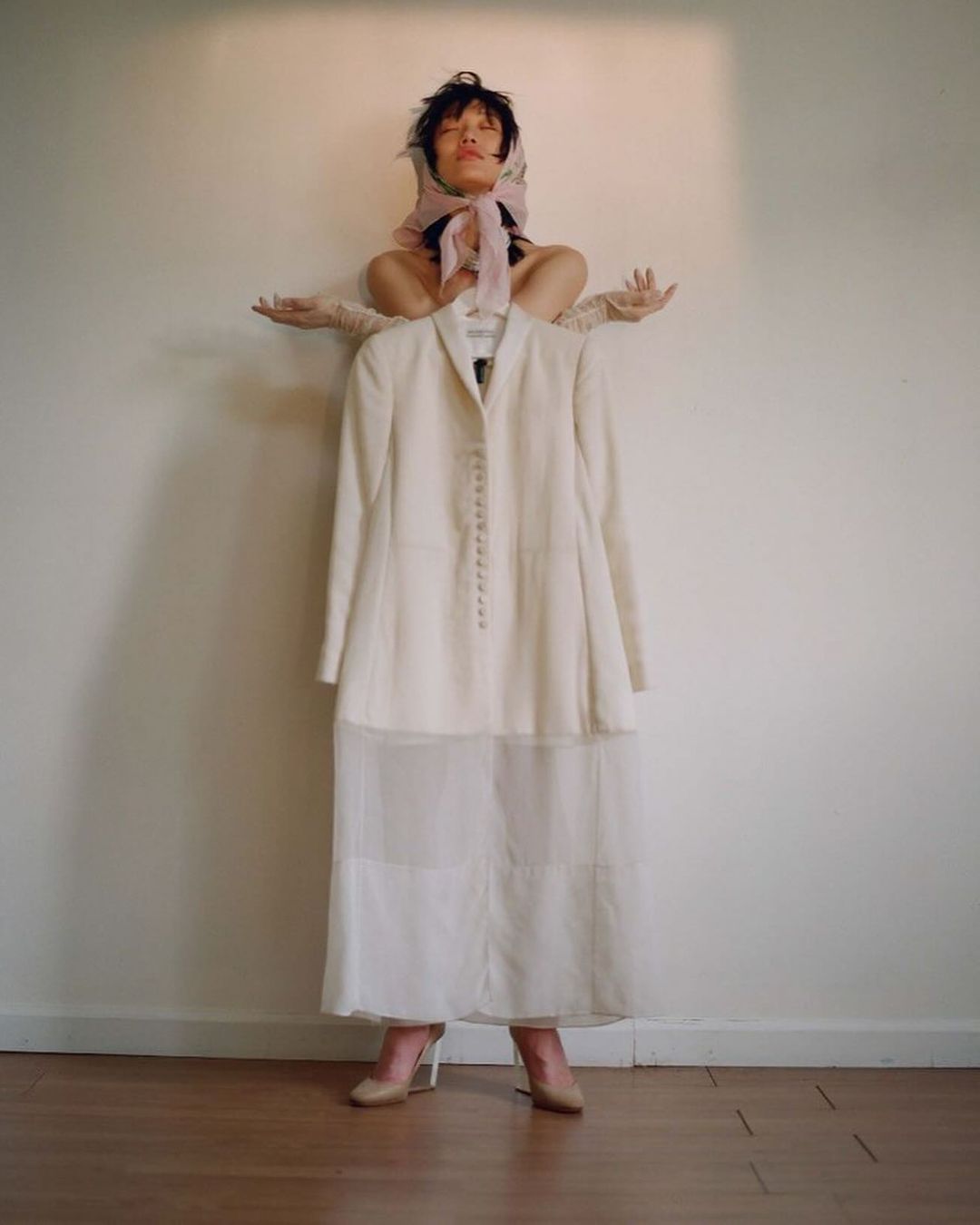 Adult woman poses with white jacket and skirt against a neutral background, wearing a headscarf and light gloves in a fashion editorial.