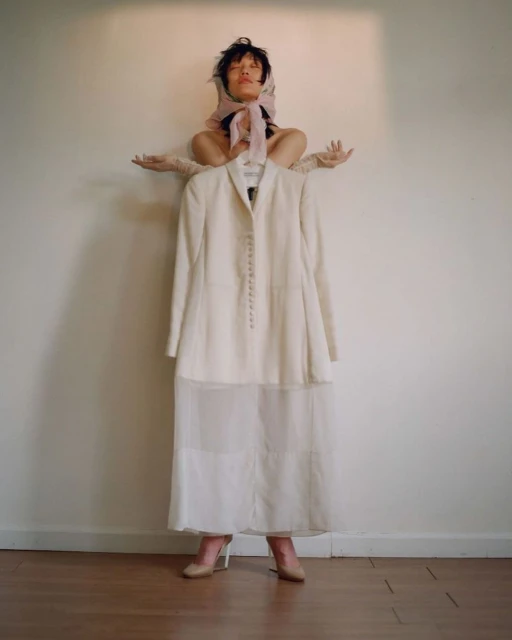 Adult woman poses with white jacket and skirt against a neutral background, wearing a headscarf and light gloves in a fashion editorial.
