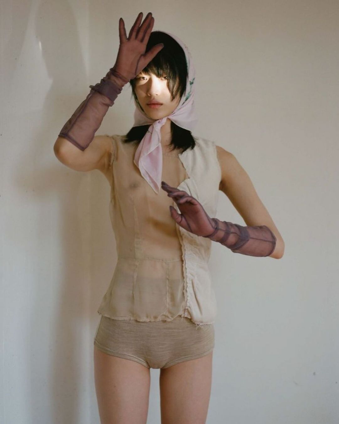 Elegant portrait of a young woman posing indoors wearing a headscarf, sheer top, knit bottoms, and sheer gloves.