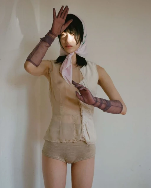 Elegant portrait of a young woman posing indoors wearing a headscarf, sheer top, knit bottoms, and sheer gloves.