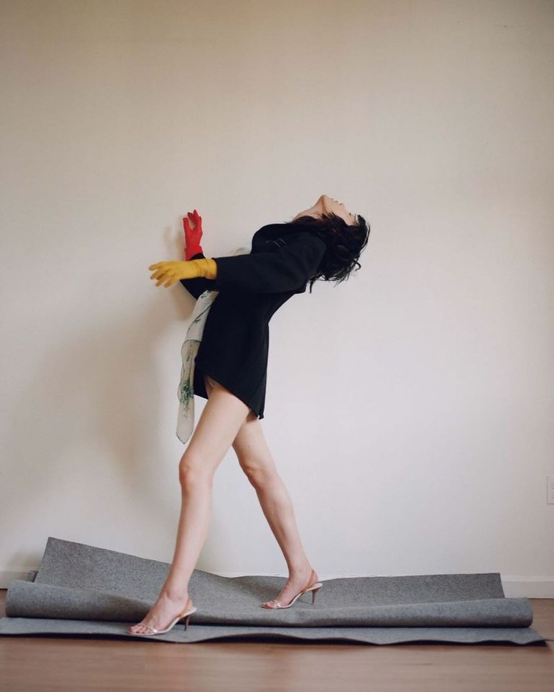 Elegant woman in a black dress posing with red and yellow gloves indoors, striking a backward arch pose, and wearing clear heels.
