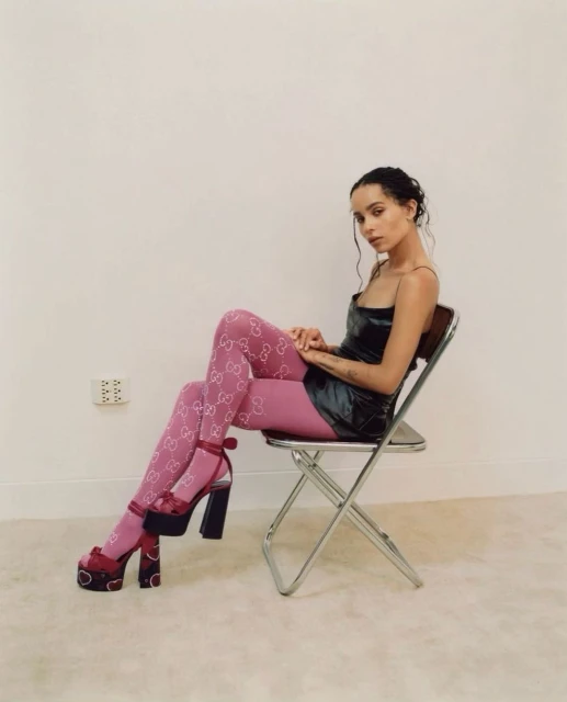 A woman with dark hair sits in a folding chair, wearing patterned pink tights, a short dress, and platform high heels against a white background.