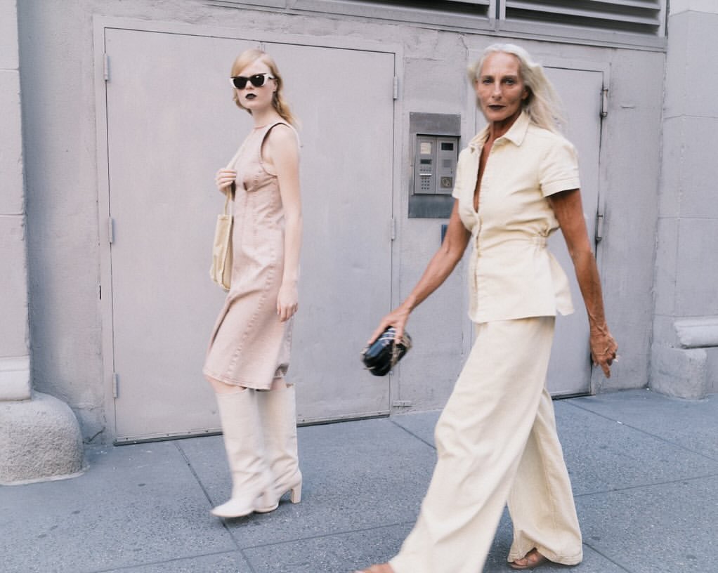 Two women in fashionable outfits walk on a city sidewalk with a building and door in the background, captured in a medium shot.
