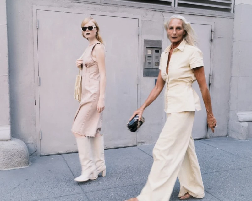 Two women in fashionable outfits walk on a city sidewalk with a building and door in the background, captured in a medium shot.