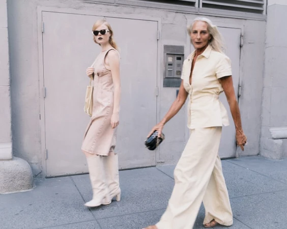 Two women in fashionable outfits walk on a city sidewalk with a building and door in the background, captured in a medium shot.