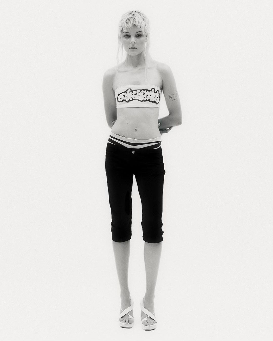 Full-length shot of a slender woman in black capri pants and a white top, posing against a bright white background in a studio.