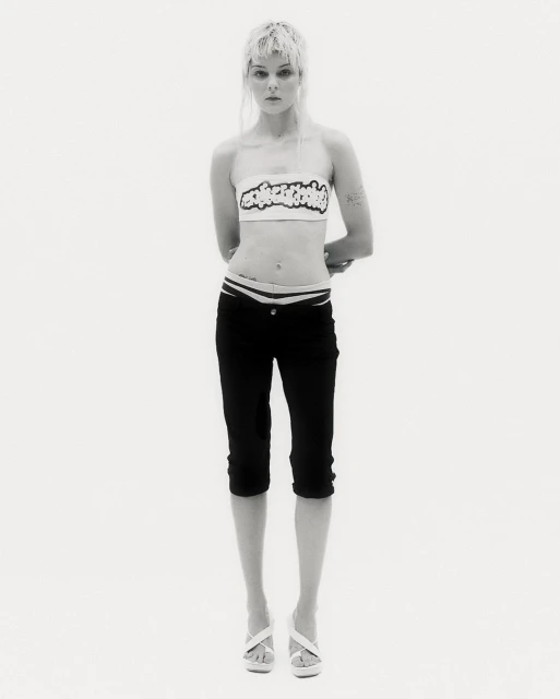 Full-length shot of a slender woman in black capri pants and a white top, posing against a bright white background in a studio.