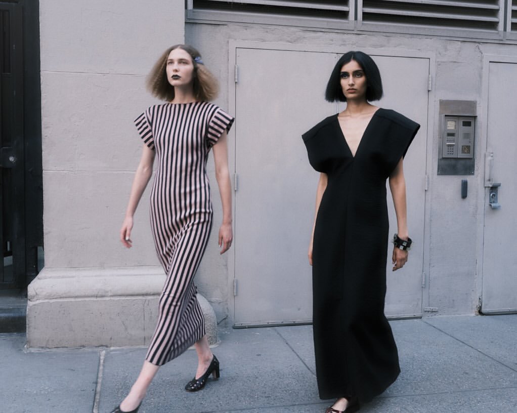 Two female models walking on a city sidewalk in front of a metal building door, wearing structured black and striped dresses.