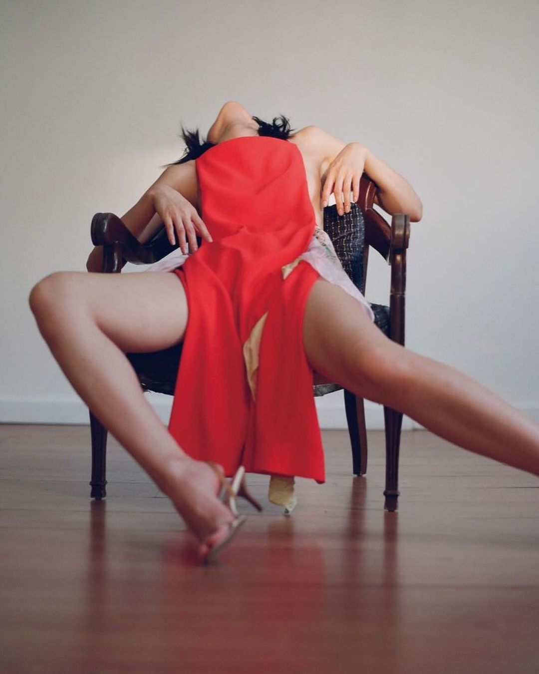 Woman reclines in a dark wooden chair, wearing red fabric and high heels, in a studio with a light beige background and wood floors.