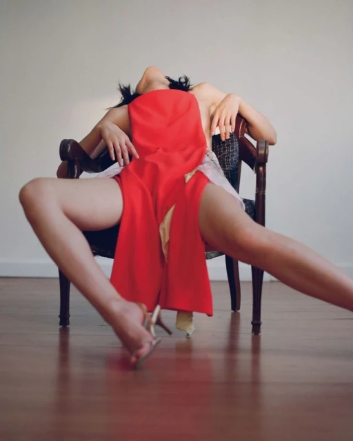 Woman reclines in a dark wooden chair, wearing red fabric and high heels, in a studio with a light beige background and wood floors.