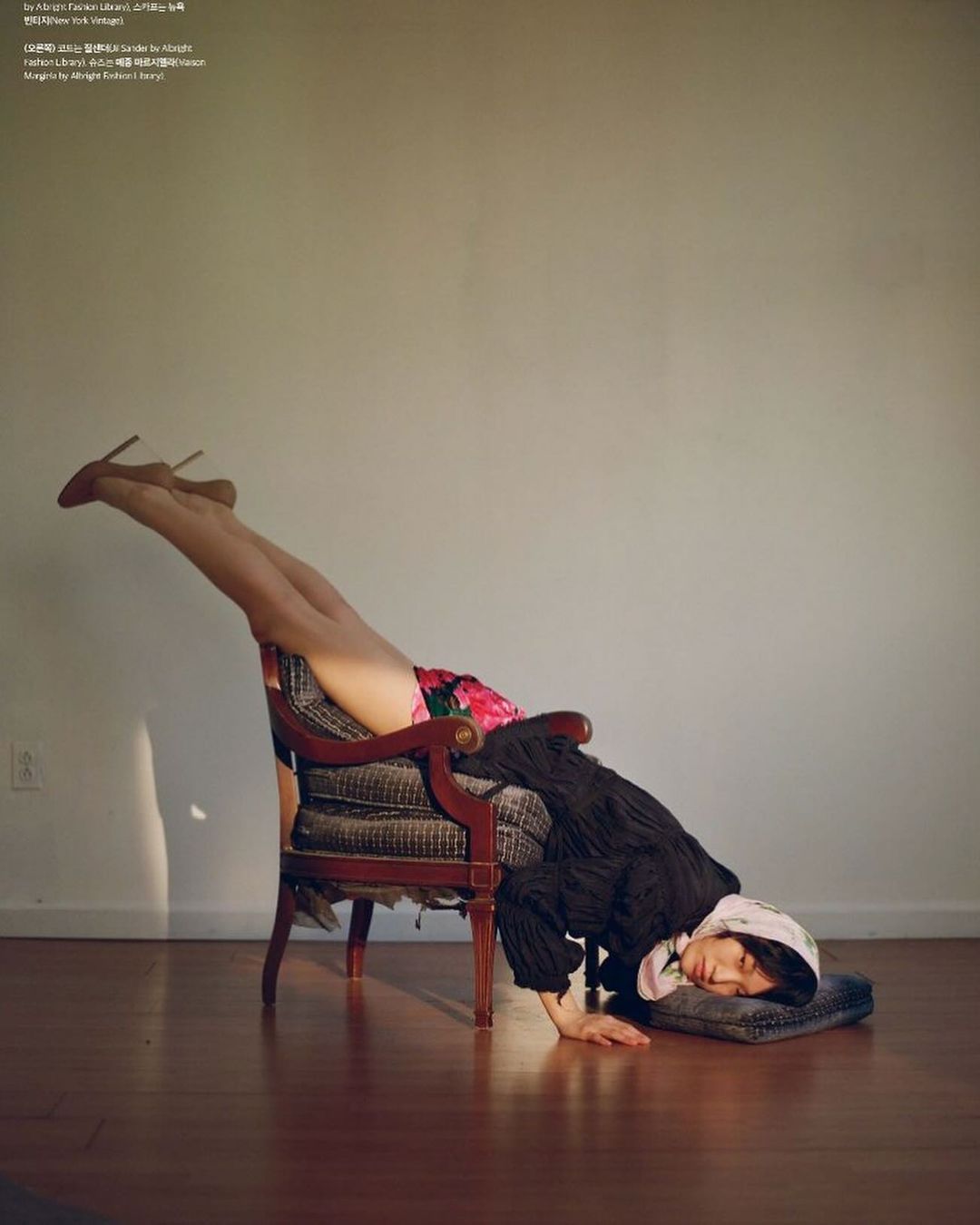 Woman in floral skirt and heels is in a handstand on an armchair, resting her head on a grey pillow on a hardwood floor.