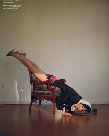 Woman in floral skirt and heels is in a handstand on an armchair, resting her head on a grey pillow on a hardwood floor.